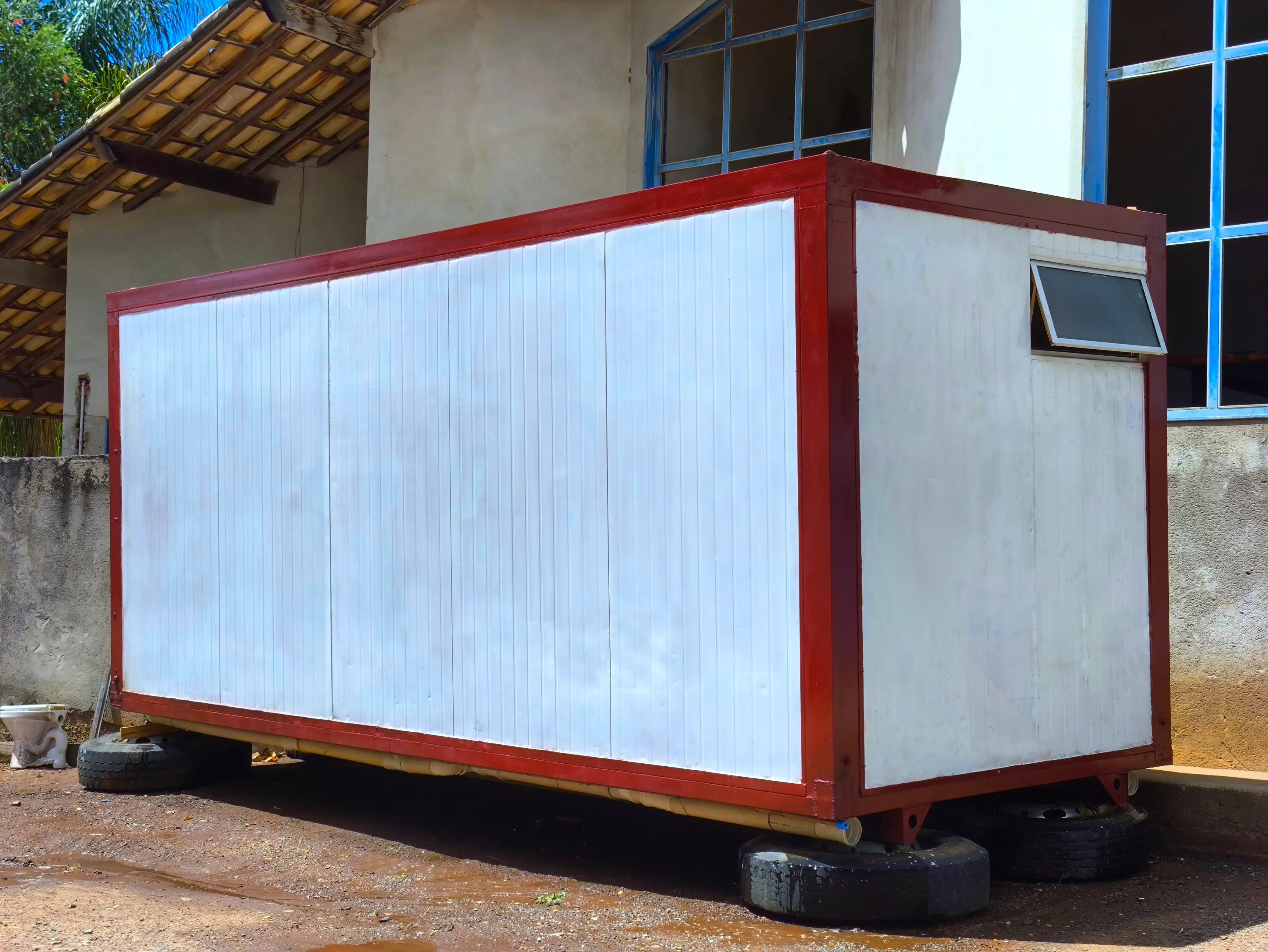 Vista externa de módulo sanitário branco com estrutura vermelha e janela basculante